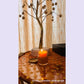 Candle in a glass holder on a decorative table with a tree-like object and curtains in the background.Actual customer photograph.