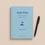 Blue book titled 'Daily Wins' with a pen on a beige background
